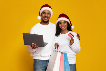 The couple wears festive Santa hats and smiles while holding a laptop and shopping bags. They engage in holiday shopping, showcasing a credit card.