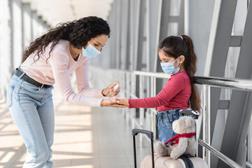 A mother applies hand sanitizer on her daughter's hands while preparing for a journey at an airport. Both wear face masks and are surrounded by modern architecture.