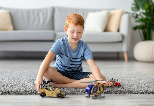 A cheerful redhead boy sits on the carpet at home, joyfully playing with toy military vehicles. He is riding a helicopter and a car, having fun after school in a bright living space.