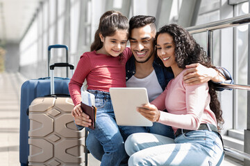 A happy family of three is sitting in an airport terminal. They are looking at a tablet together, sharing smiles and laughter. Their luggage is nearby, showing they are traveling.