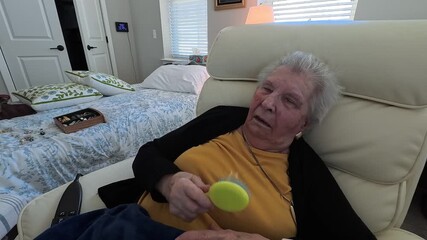 An elderly woman brushes her hair, sitting in a comfortable chair in a warm nursing home atmosphere