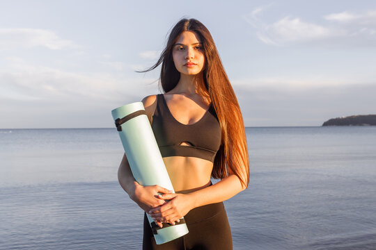 Woman holding yoga mat by the sea at sunset, fitness lifestyle, yoga training, outdoor wellness, healthy routine, seaside workout, calm evening yoga vibes.