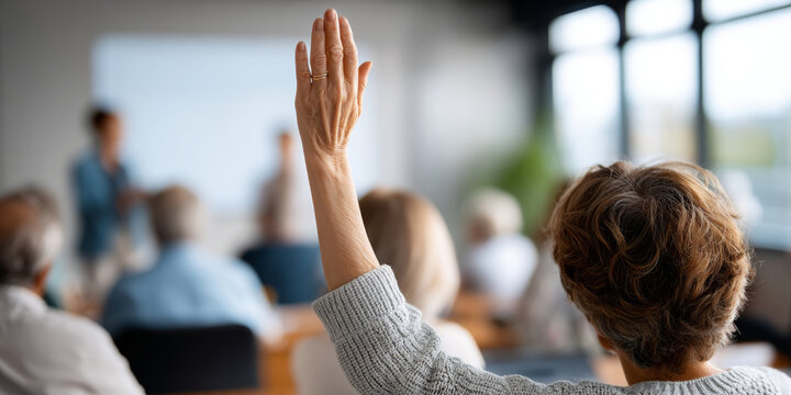 Senior woman raising hand in classroom setting during educational seminar or workshop with blurred instructor and audience in background