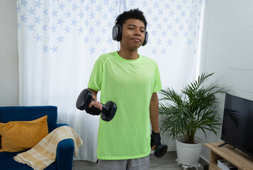Brown skinned teenager lifting dumbbells at home, listening to music with wireless headphones and enjoying his workout. Concept of modern home training