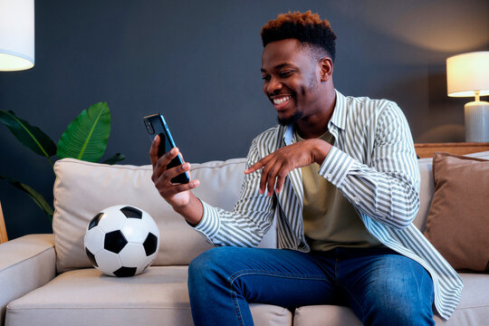Young adult Black man sitting on sofa watching TV while holding smartphone and smiling, soccer ball placed beside him, engaging with content related to soccer match on television