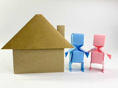 A blue and pink origami couple stands next to a cardboard kraft house, representing joint homeownership, family, or real estate concepts on a white background.