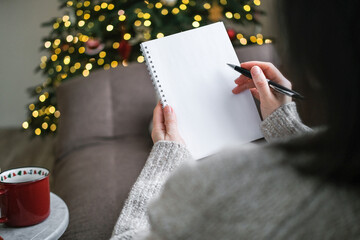 Over-the-shoulder view of person in sweater writing in blank spiral notebook with pen with blurry...
