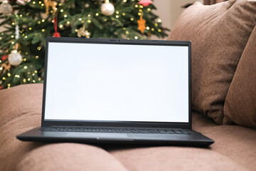 Laptop with blank white screen mockup is placed on cozy brown sofa with decorated Christmas tree in blurred background. Holiday websites, online shopping, remote work during festive season