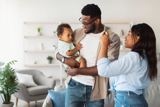In a cozy living room, joyful black parents interact with their baby. The father holds the infant while the mother shows toys. The atmosphere is filled with laughter and love.