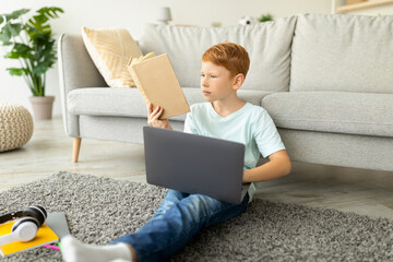 A young redhead boy sits on the floor of his room, focused on homework while holding a notebook on his lap. He uses a laptop to assist in his studies, immersed in learning at home.
