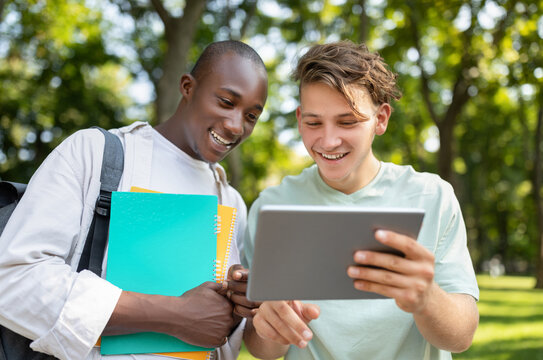 Two young men are enjoying a lively moment in a park as they look at a tablet together. They are surrounded by green trees and bright sunlight, creating a cheerful atmosphere. - Powered by Adobe