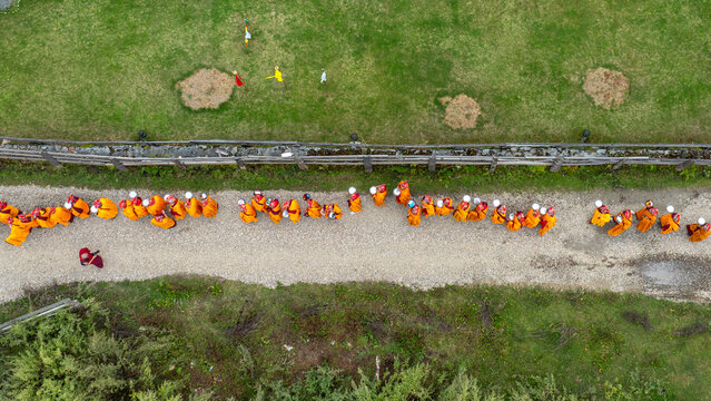 Aerial view of monks in vibrant orange robes line a pale path amidst lush green fields, Phobjikha, Wangdue Phodrang, Bhutan.