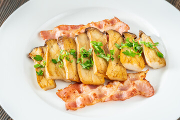 Fried eryngii mushrooms served with crispy bacon slices on white plate