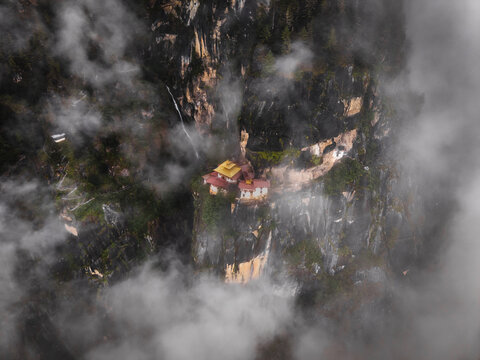Aerial view of a golden-roofed temple perched precariously on a sheer cliff face, shrouded in ethereal mist and cascading waterfalls, Tigers Nest, Paro, Bhutan.