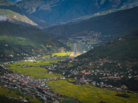 Aerial view of the Paro Airport runway cutting through the lush green valley, surrounded by traditional Bhutanese architecture, framed by towering mountains, Paro, Bhutan.