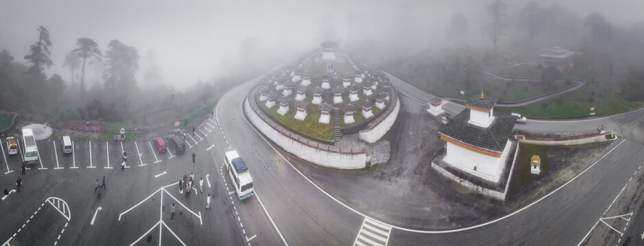 Aerial panoramic view of the Dochula Chorten's white stupas stand amidst the dense fog, complemented by the geometric precision of the parking lot, Hungtso, Bhutan.