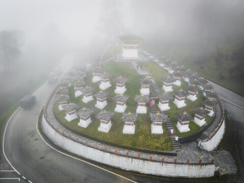 Aerial view of the Dochula Chorten shrouded in mist, its white stupas standing out against the green grass and winding road, Hungtso, Bhutan.