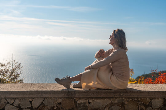 Woman Ocean View Relaxation: Coastal autumn serenity; mature woman sits cliffside enjoying panoramic ocean vista.