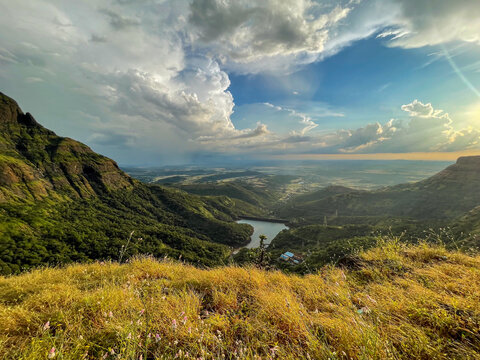 Breathtaking view from Konkan Kada in Bhandardara with lush green mountains, golden grass, dramatic monsoon clouds, and a serene valley reservoir in Maharashtra, India.