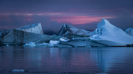 Arctic icebergs float in calm water under a dramatic pink and purple twilight sky.