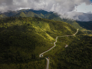 Aerial view of a serpentine road winding through verdant hills and shadowed valleys beneath a canopy of swirling clouds, Black Mountain range, Wangdue Phodrang, Bhutan.