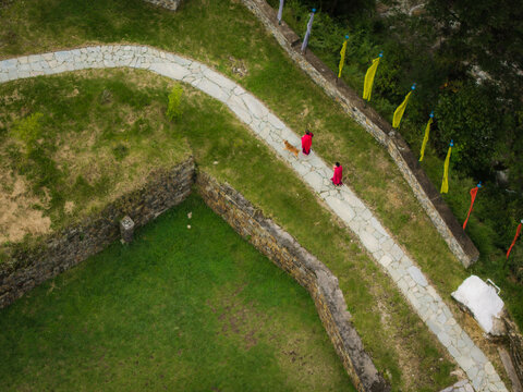 Aerial view of two monks in red robes walking along a winding stone path near Chendebji Chorten Stupa, framed by stone walls and colorful flags, Chendebji, Trongsa, Bhutan.