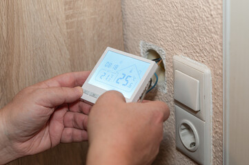 Man installing a digital underfloor-heating thermostat, connecting the device to wall wiring as the display shows temperature settings and controls.