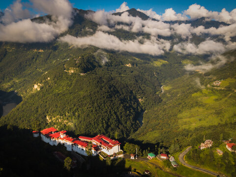 Aerial view of a fortress-like structure with red roofs nestled amidst the rolling green hills and cloud-capped mountains, Trongsa, Trongsa, Bhutan.