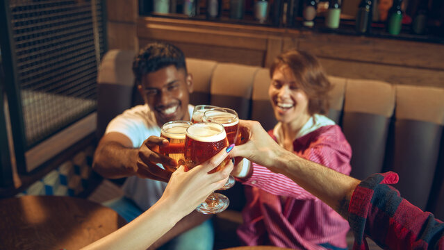 Friends clinking beer glasses with joyful expressions in pub. Concept of festive nightlife imagery, beer brand storytelling, weekend entertainment, social bonding, and friendly celebration moments.