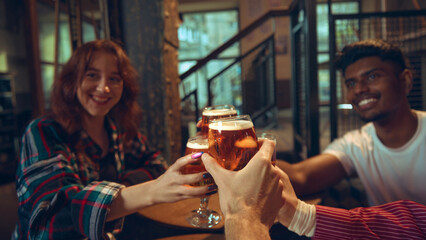 Group raising beer glasses around pub table in warm interior. Concept of brewery marketing,...