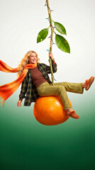 Woman Joyfully Swings on Giant Orange with Festive Lights and Vibrant Green Outfit. Winter, Christmas or New Year Concept