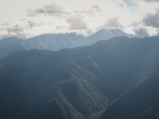 Aerial view of undulating mountain ranges veiled in a soft, blue-gray haze, where peaks meet the cloudy sky in a serene dance of light and shadow, Paro, Paro, Bhutan.
