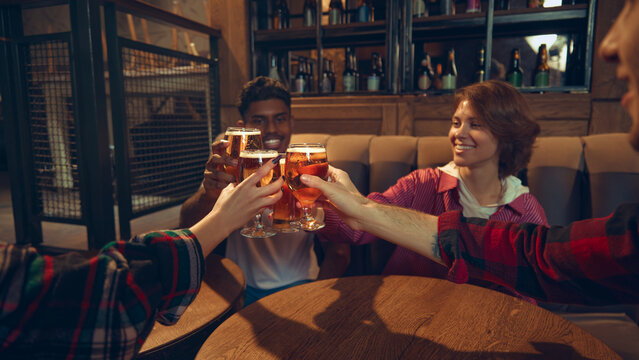 Friends clinking beer glasses while sitting together in cozy pub. Concept of bar events, brewery visuals, social advertising, nightlife promotions, and beverage-centered lifestyle marketing. - Powered by Adobe