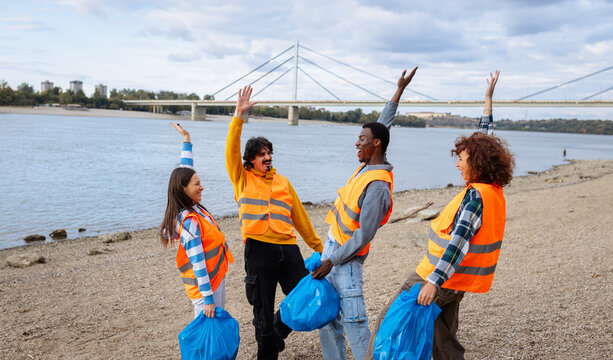 Portrait of a group of volunteers standing on the riverbank celebrating with their hands up after collecting plastic waste.