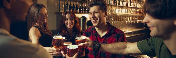 Friends clinking beer glasses at pub counter with warm evening lighting. Concept of beverage promotion, nightlife visuals, bar marketing, social leisure. Banner