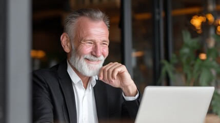 Happy senior man with gray beard working on laptop in modern cafe, smiling with warmth and contentment in soft light