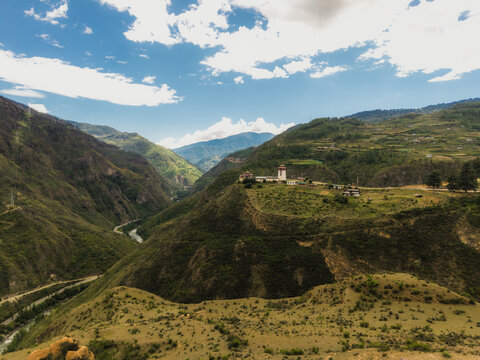 Aerial view of the stark white architecture of a distant temple perched atop a lush green hill, contrasting against the deep valley, Raga, Paro, Bhutan.