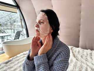 Adult brunette woman applying a sheet face mask at home, adjusting skincare mask for moisturizing and anti-aging treatment, self care routine in a cozy domestic interior