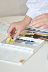 A close-up of a hand picking a yellow watercolor block from a paint set, with brushes and a palette visible. Creative, colorful, and cozy mood suited for art, hobbies, and craft projects.
