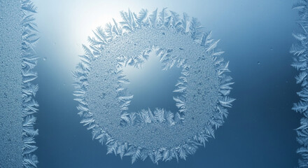 Closeup of intricate frost patterns forming a circular shape with a stylized letter or symbol on a cold window pane against bright sunlight