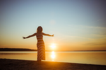 Fototapeta premium Silhouette of young sexy woman in full-length stands with an arms are outstretched against the sunset on the lake's shore in long striped dress. Greeting to the sun. Women's freedom. Summer vibe.
