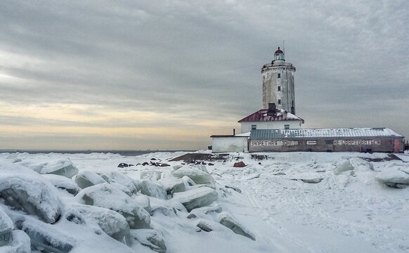 Ice ridges and a snowy field in front of the Tolbukhin Lighthouse on the frozen Gulf of Finland, with keeper&rsquo;s buildings, a Russian warning sign, and a dark water channel on the horizon.