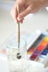 A close-up of a hand gripping a wooden watercolor brush as it dips into a glass of water, with a colorful palette in the background, capturing creative process and artistic focus.