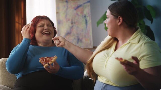 Two curvy women eating pizza and singing into imaginary microphone, fun at home