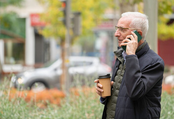 Senior man communicating on his cell phone and holding a coffee cup outdoors