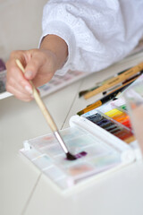A woman in a white sleeve paints with a brush on a watercolor palette, surrounded by colorful tubes. The scene captures creativity, focus, and hands-on art play.