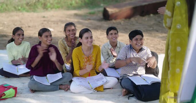 Indian happy girl boy sitting on mat clap hand teacher tutor share good news have fun joy rural area ground place. Teen age child group talk do study book enjoy desi life open air class field new day