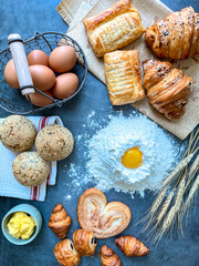 Top-down flat lay composition of a bakery workspace featuring a central mound of flour with a raw egg yolk, surrounded by fresh croissants, sesame buns, puff pastries, a basket of brown eggs, butter, 