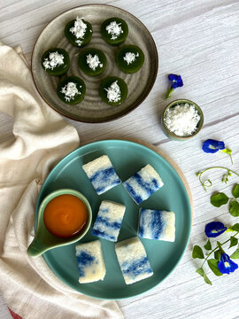 High-angle view of assorted traditional Nyonya Kuih, featuring green Kuih Kaswi topped with grated coconut and blue Pulut Tai Tai glutinous rice cakes served with Kaya jam on a wooden table.