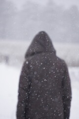 Płatki śniegu wirują w powietrzu, a w tle rozmazana kobieta w ciemnym płaszczu idzie w stronę lasu. Zimowa sceneria nabiera dynamiki i tajemniczego klimatu.